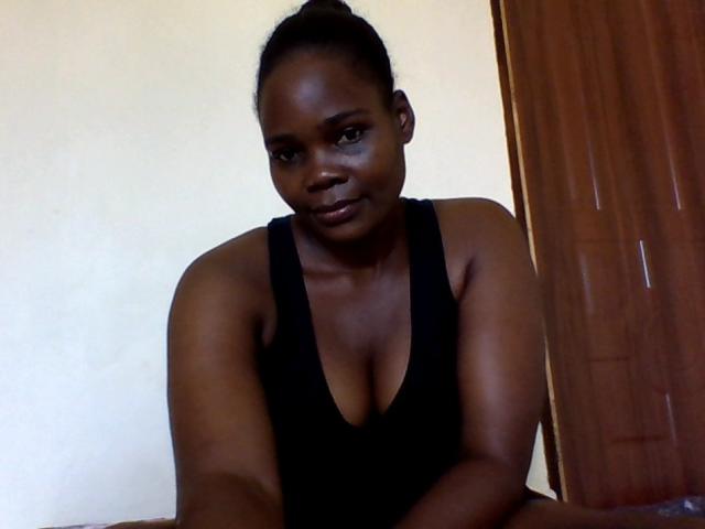 MedeyaX with dark skin, black hair in a bun, wearing a black tank top showing her boobs, sitting with a slight smile, arms resting on a surface.