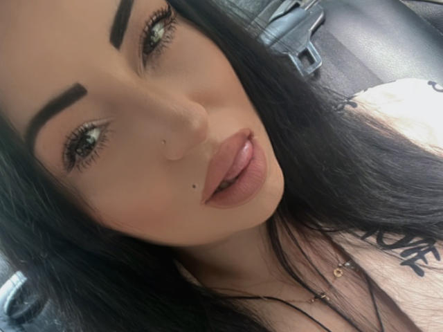 SmileAndrea has long black hair, thick eyebrows, green eyes, glossy pink lips, and a small beauty mark under her right lip. She's wearing a necklace with a gold cross.