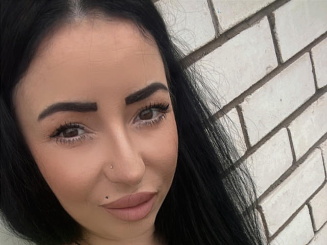 SmileAndrea has black hair, brown eyebrows, long eyelashes, and two facial piercings—one on her nose and one below her lips. Her skin is light, and she's smiling.