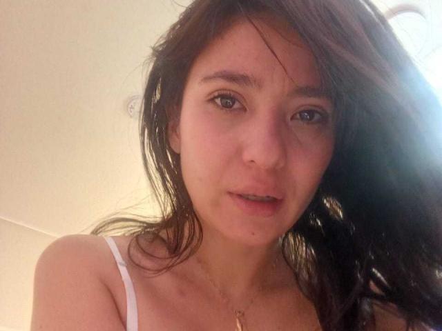 CattsyCathy has a cute face with brown eyes, slightly messy long dark brown hair, and wears a white spaghetti strap top showing her small tits and a gold necklace.