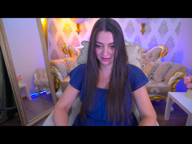KarinaClover, con lunghi capelli castani, un abito blu e rossetto rosa, seduta su una sfarzosa poltrona, sorridente, in una stanza illuminata in modo caldo, con uno specchio con cornice dorata..