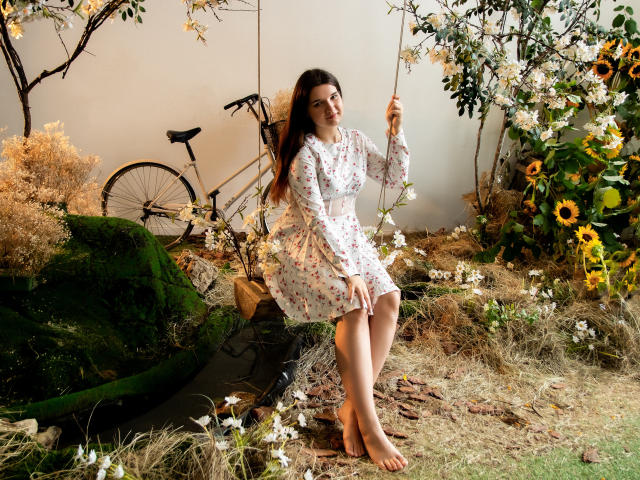 BonnieFlower sentada descalza sobre una roca, vestida con un vestido floral, sosteniendo un tallo de flor, una bicicleta al fondo, rodeada de flores y hierba seca, cabello castaño largo..