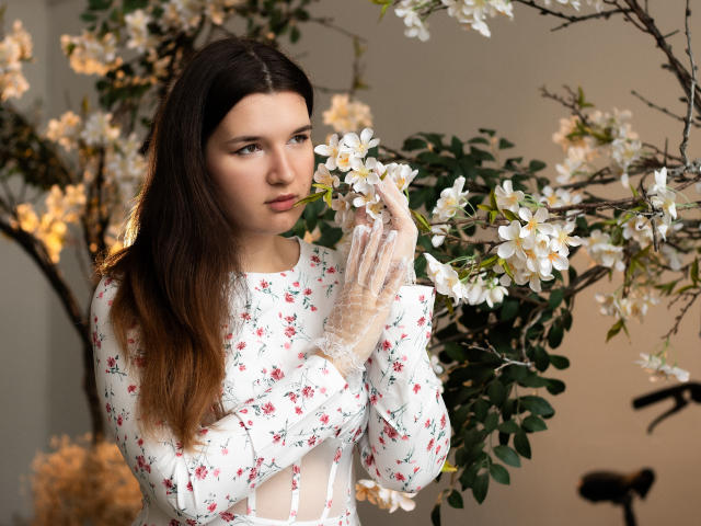 BonnieFlower, con cabello largo y castaño, vestida con un vestido blanco con estampado floral, sosteniendo flores blancas, usando guantes translúcidos, mirando a la cámara con una sonrisa sutil, de pie frente a ramas con flores..
