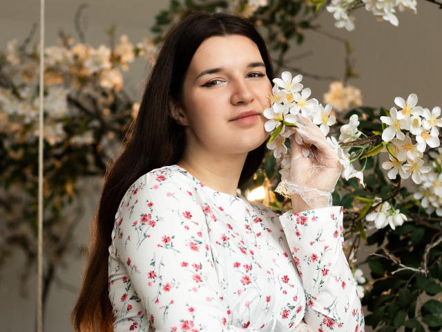 BonnieFlower with long brown hair, wearing a white floral dress, holding white flowers to her nose, smiling softly, wearing lace gloves, looking cute and relaxed.