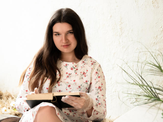 BonnieFlower, con el pelo largo y castaño, vestida con un vestido floral, sostiene un libro con una cubierta negra, lleva guantes transparentes y está sentada con una expresión pensativa..