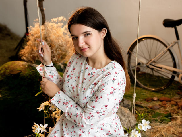 BonnieFlower, con un busto de tamaño medio, vestida con un vestido blanco con estampado floral, sentada en un columpio con flores, cabello castaño, una ligera sonrisa, manos en las cuerdas del columpio, una bicicleta en el fondo..