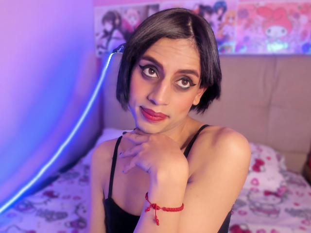EimyFlorian with short black hair, dark eyeliner, and red lipstick, wearing a black spaghetti-strap top, looking up with a hand near her chin, red bracelet on her wrist.
