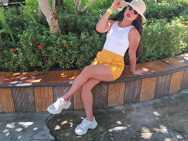CarolineWill with long brown hair, white tank top, yellow shorts, white sneakers, and straw hat, sitting on wooden wall, holding hat, showing her ass, and wearing red lipstick.