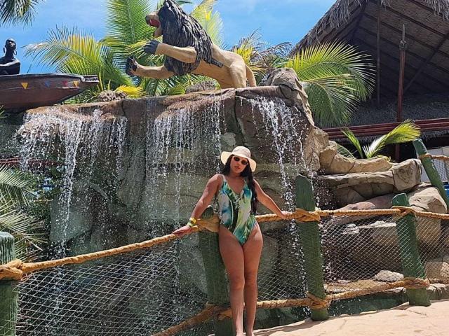 CarolineWill with tan skin and dark hair, wearing a sunhat, green and blue patterned one-piece, standing under a rock waterfall, holding a bamboo fence. Water cascades down.