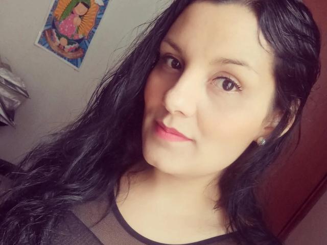 ScarlettMancini has wavy black hair, light skin, and pink lipstick, wearing a sheer black top, with small earrings, and a slight smile, in a casual selfie.