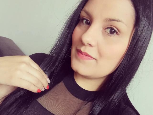 Scarlett Mancini, met lang zwart haar, lichte huid en rode lippenstift, draagt een zwarte, doorschijnende top, toont rode nagellak en heeft een subtiele glimlach, terwijl ze naar de camera kijkt..