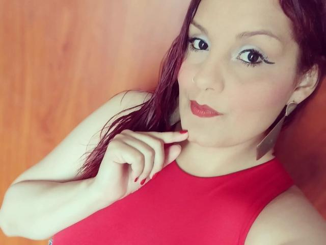 ScarlettMancini has pale skin, red lipstick, and wet red hair, wearing a red sleeveless top, with red nails touching her chin, and blue eyeliner.