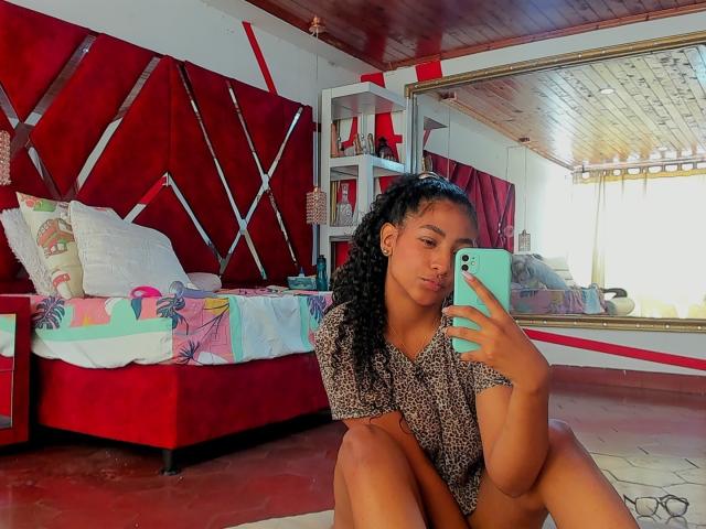 NatalyBlonder with curly black hair, brown skin, and small tits, squatting in a room, taking a selfie with a turquoise phone, wearing a leopard print shirt, and red bed.