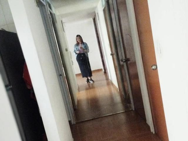 ScarlettMancini with long brown hair, wearing a blue shirt and dark skirt, standing in a hallway, holding a phone, and wearing black sneakers, looking down at it.