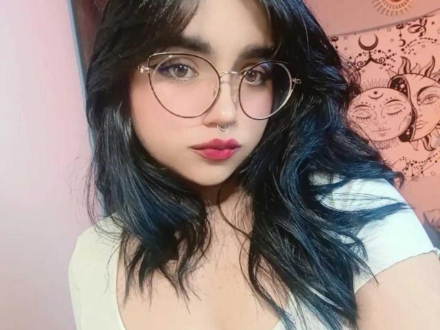 LovelyLilith has black wavy hair, glasses, pink lipstick, nose ring, and a white top, showing her tits in a selfie with a sexy, cute look.