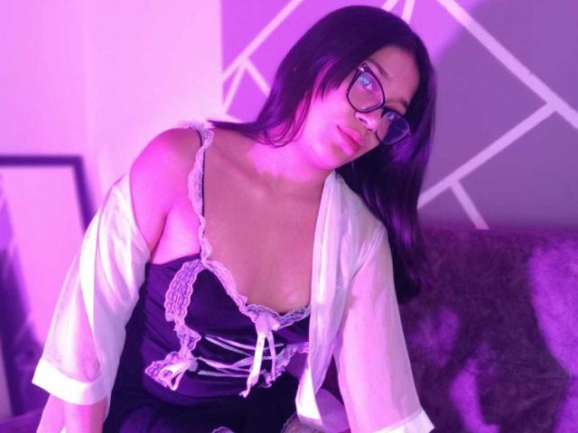 CuteVioletta with black hair and glasses, wearing a black dress with white lace, and a white shirt off her shoulders, purple lighting, looking slightly to the side.
