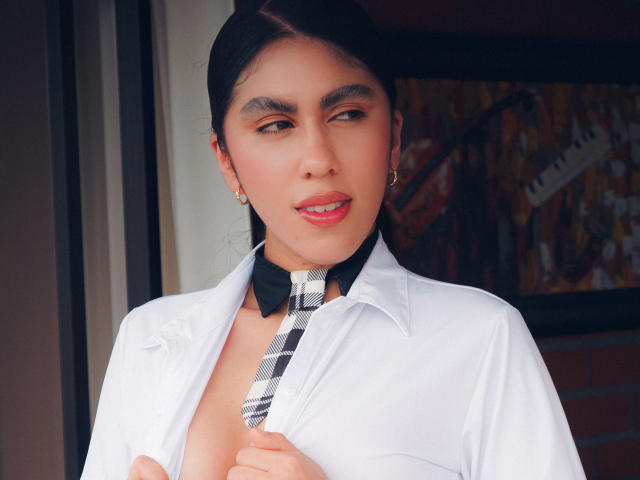 LilyGreay with dark hair, light skin, and red lipstick, wearing a white shirt and black/white tie, holding tie, showing cleavage, small earrings, looking slightly to the side.