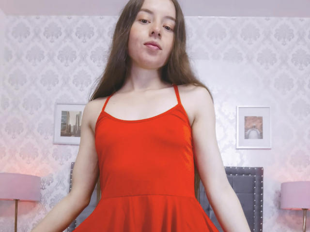 VioletaNova, with long brown hair, wearing a red dress, has small tits and a cute face, standing in a room with pink lamps and patterned wallpaper.