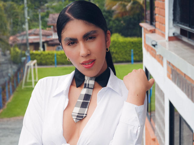 LilyGreay with black hair, white shirt, black and white checkered tie, and gold earrings, holding shirt collar, smiling, red lipstick, small gold hoop earrings, and a cute face.
