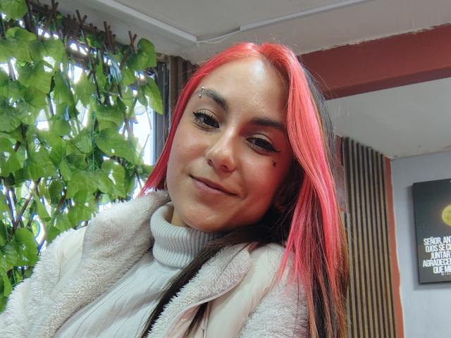 AmyStreetwise69 with red and black hair, black eyeliner, and a white turtleneck, smiling slightly, wearing a fluffy white coat, small black mole on her right cheek.