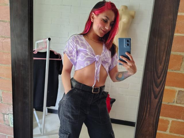AmyStreetwise69 with pink hair, tied-up purple top showing her tits, black pants, tattoo on left arm, smiling mirror selfie, hands in pockets, brick wall, mirror reflection.