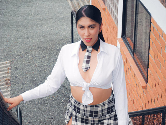 LilyGreay with tied white shirt exposing her tits, black-and-white plaid skirt, matching necktie, and black pigtails, standing outdoors, hand on brick wall, looking at camera.