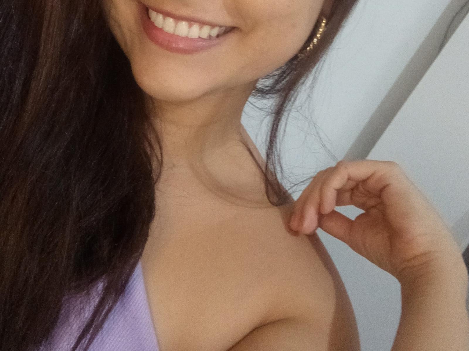 RebecaLovely with long brown hair, smiling, wearing a purple top, showing her bare shoulder and upper chest, holding her hair, gold earrings, and her hand near her collarbone.