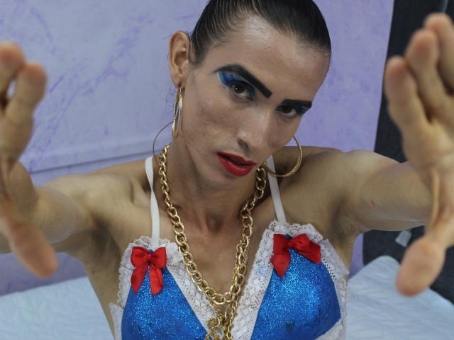 ShantalaNastasiia with blue glitter bra, red bows, white lace, gold chain, hoop earrings, and red lipstick, reaching forward, dark blue eyeshadow, slicked-back hair, looking intense.