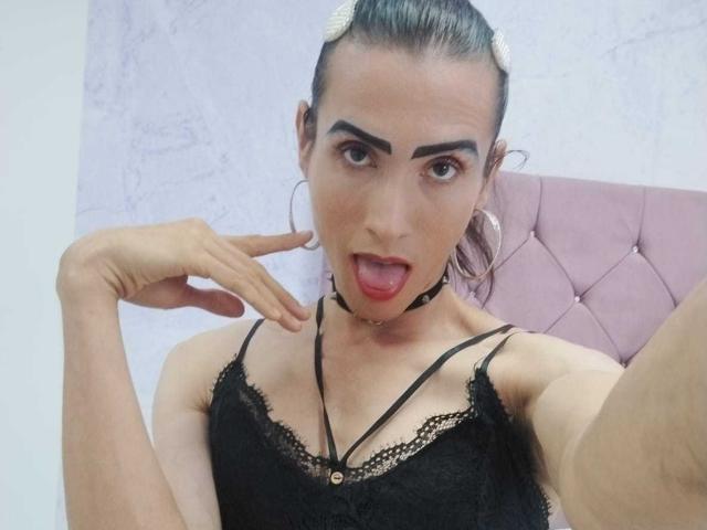 ShantalaNastasiia with dark hair in a bun, black lace top, choker, and hoop earrings, sticking out tongue, pointing with index finger, and showing off small tits.