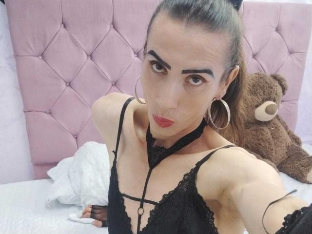 ShantalaNastasiia with dark eyebrows, red lipstick, black lace top, hoop earrings, and arm raised, showing a slim, fair-skinned, flat-chested, androgynous upper body. Brown teddy bear in background.