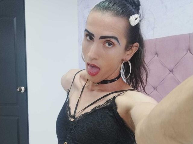 ShantalaNastasiia with dark hair in a ponytail, wearing a black lace top, choker, and hoop earrings, sticking out her tongue, with eyebrows raised. Her tits are small.