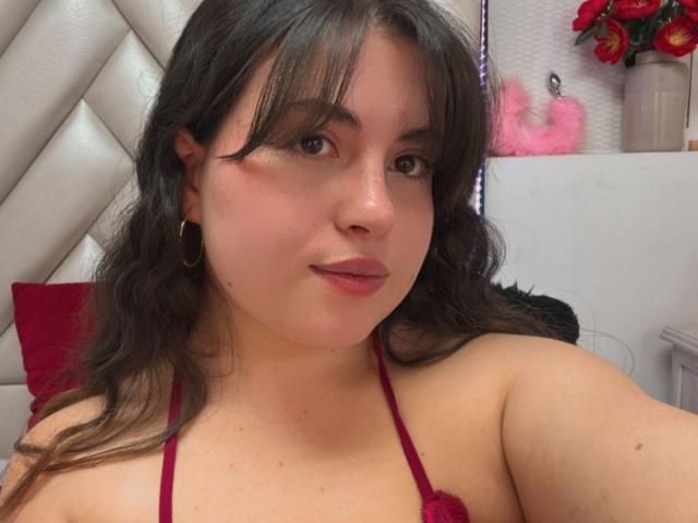 VicluX has medium brown wavy hair, wearing a red halter top, small gold hoop earrings, and a slight smile, with visible pink lipstick and a soft, fair complexion.