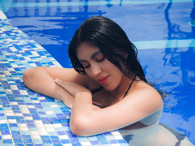 LilyGreay with wet black hair, arms folded over blue tiled pool edge, eyes closed, red lipstick, water droplets on pale skin, smiling softly, topless, in bright blue pool.