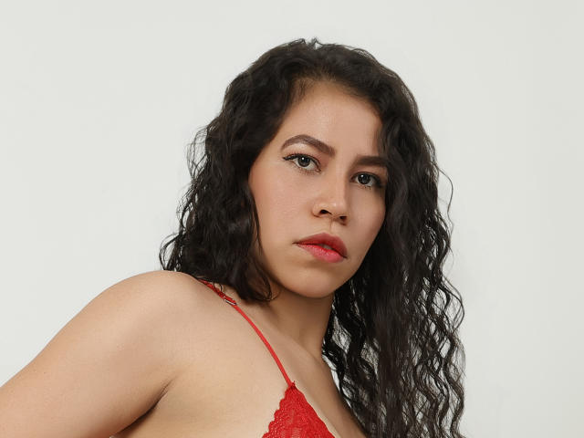 AlessandraValmont with wavy black hair, red lace bra, and serious expression, showing her tits against a plain white background. She has light brown skin and red lipstick.