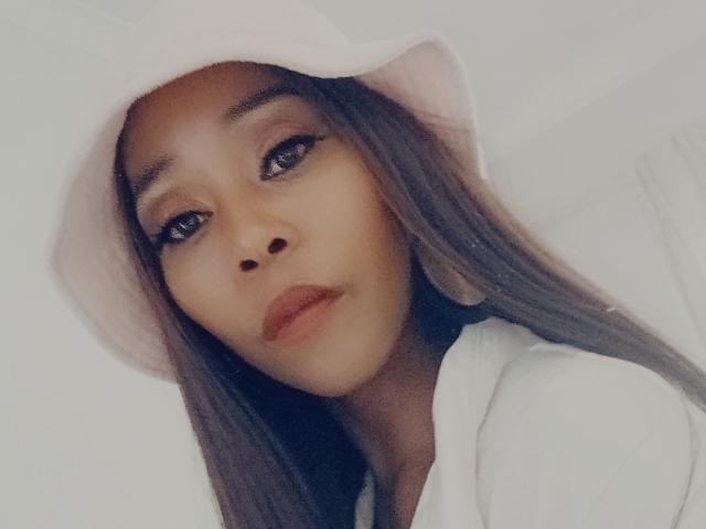 BuffyX mit langen, braunen Haaren, die einen rosa Eimerhut und einen weißen Umhang trägt, blickt in die Kamera mit geschürzten Lippen und dunklem Eyeliner. Weiches, leicht verschwommenes, feminines Gesicht..