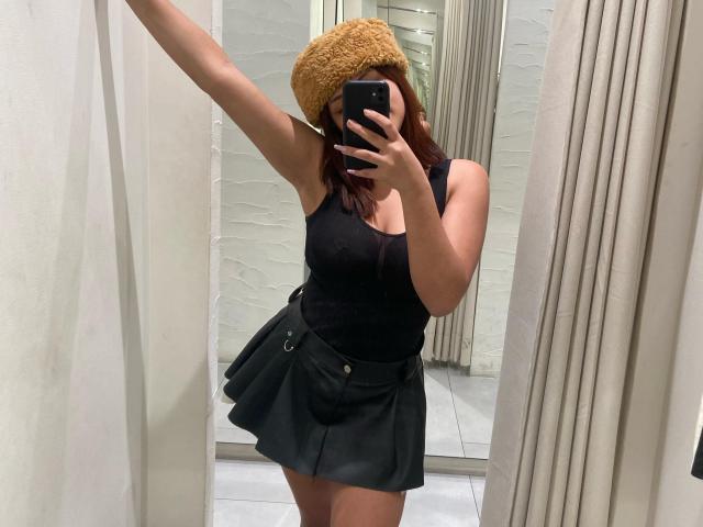 KhloeRose with brown hair, wearing a yellow knit hat, black tank top, and a black pleated skirt, taking a mirror selfie in a changing room. Boobs visible.