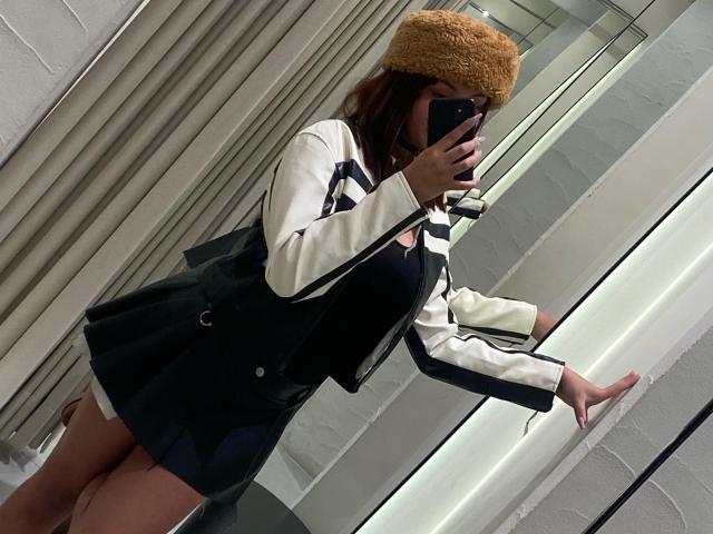 KhloeRose with tan fur hat, black dress, white and black jacket, holding phone, mirror selfie, black purse, black skirt, showing her bare ass, standing, reflective background.