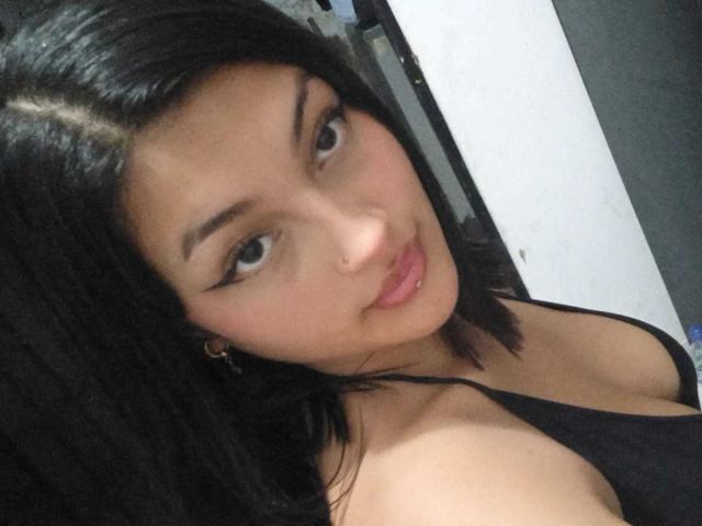 KhloeRose with black hair, looking at the camera, wearing a black top, showing her tits, pink lips, small nose, brown eyes, and gold earrings.