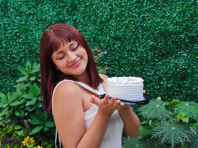KhloeRose with red hair, white dress, holding a white-frosted cake in front of her, smiling with eyes closed. She's got a cute ass and small tits.