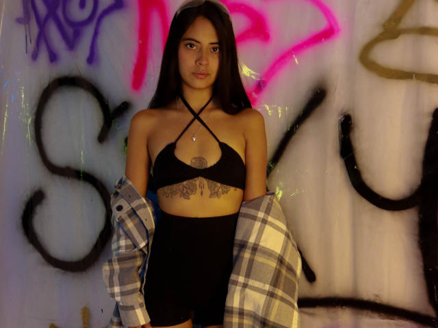 TamaraPeach with long dark hair, black criss-cross bikini top, black shorts, plaid shirt off, tattoos under breasts, standing, graffiti background, serious expression, medium skin, small breasts.