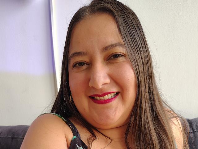 AgathdTeheran has a chubby face with brown eyes, long brown hair, and red lipstick, smiling at the camera with her nose slightly upturned. She's wearing a black top.