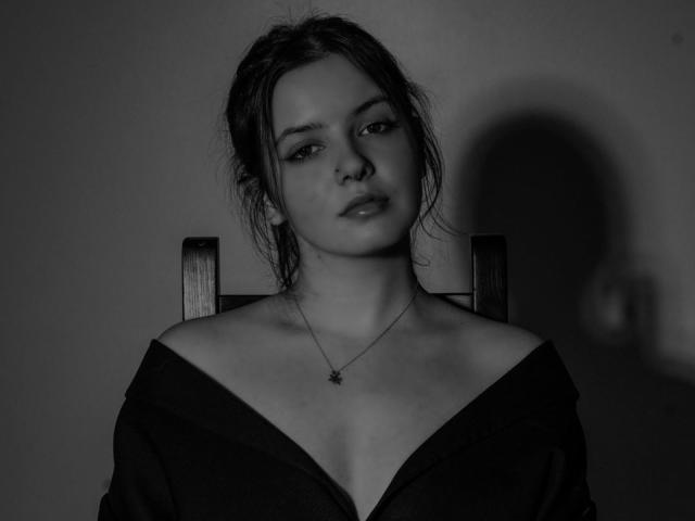 Anna : Élégante, avec des cheveux noirs coiffés de manière désordonnée, portant un haut noir à une épaule, un collier avec un pendentif en forme d'étoile, assise sur une chaise, regardant la caméra, en noir et blanc..