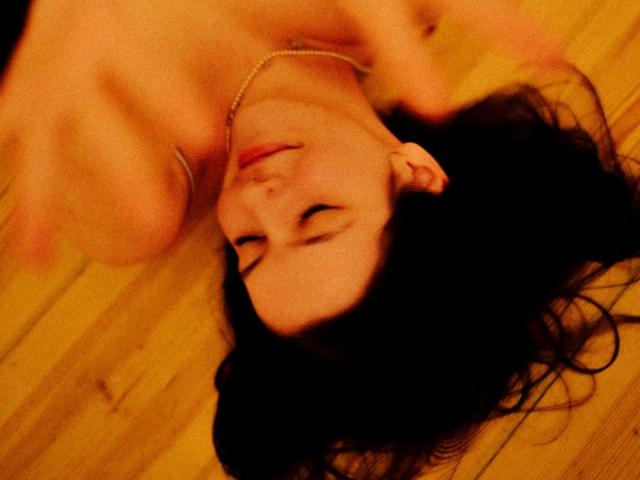 AnnaGlamorous liegt auf einem Holzboden, oben ohne, mit offenem, dunklem Haar, geschlossenen Augen und einer Halskette. Ihre nackten Brüste sind sichtbar, und ihr Gesicht ist entspannt..