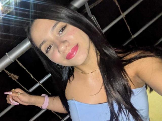 AshlyDubois has long black hair, pink lipstick, and wears a strapless blue top, showing off her small tits and a pink wristband, smiling at the camera.