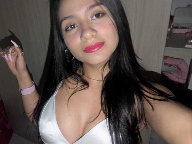 AshlyDubois with long black hair, pink lipstick, white tank top showing her tits, holding a phone, pink watch, gold ring, and hoop earrings, smiling slightly.