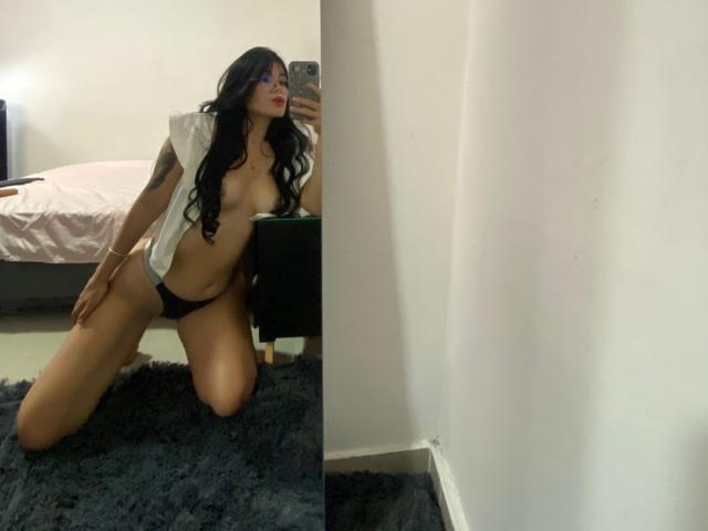 JuliianaSmiitt kneeling on a rug, wearing black panties and a white shirt open to show her tits, taking a mirror selfie with long black hair and red lipstick.