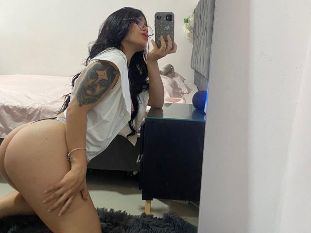 JuliianaSmiitt with long black hair, glasses, white shirt, and grey thong, leaning forward, showing her big ass and tattooed arm, taking a mirror selfie with a phone.