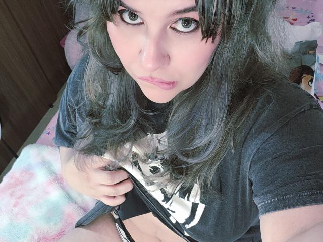 Akkanee with grey hair, blue eyes, and pink lips, wearing a black shirt, lifting it to reveal her flat stomach and black underwear, sitting on a pink blanket.