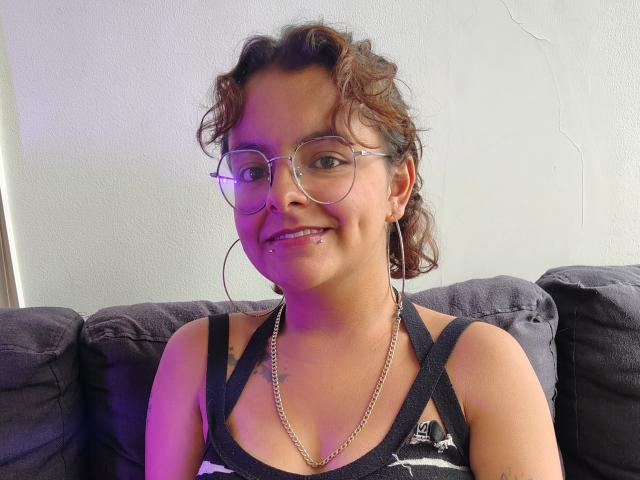 DarcyTear with curly brown hair, glasses, black tank top, gold chain, nose ring, and lip piercing, smiling on a dark couch. Purple light on her face.