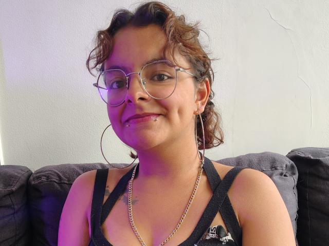 DarcyTear with curly brown hair, glasses, hoop earrings, and a black tank top, smiling with a small lip piercing, showing her tits, sitting on a dark couch.